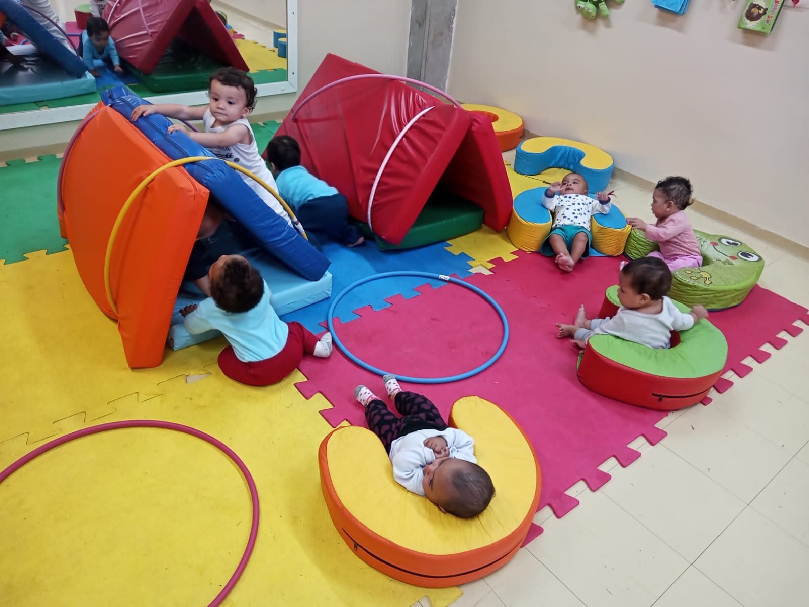 Bebês brincando de cabana com colchonetes e bambolês.